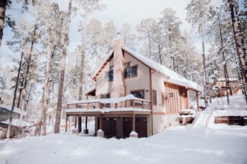 A home with off-white siding and orange trim in a snowy yard surrounded by tall pine trees.