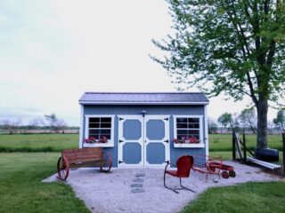A small blue garden shed with white doors and window frames. Two benches and a chair sit in front.