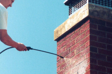 A person in a white shirt pressure washes a red brick chimney with a silver chimney cap.