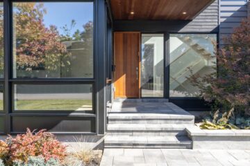 The picture depicts the front entryway of a house that is primarily made of glass. The door is cedarwood and there is a stone walkway.
