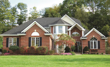 A red brick, two story home with white trim and black shutters. The front of the home is landscaped with bushes and a small tree by the front door.