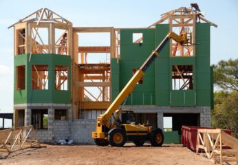 An image of a home being built is pictured during the day. There is a person standing on the building frame.