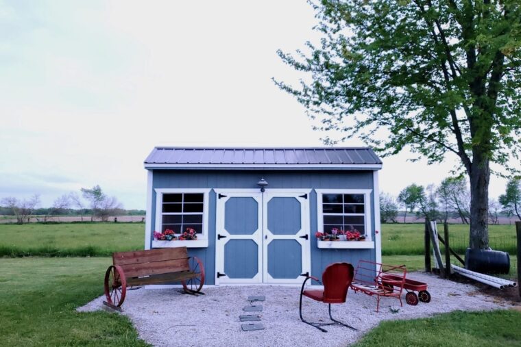 A small blue garden shed with white doors and window frames. Two benches and a chair sit in front.