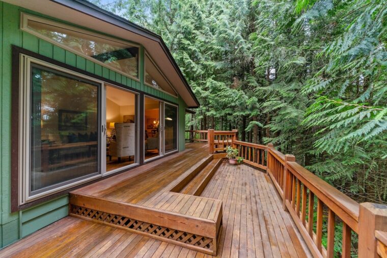 A home with green siding and a wooden deck surrounded by pine trees.
