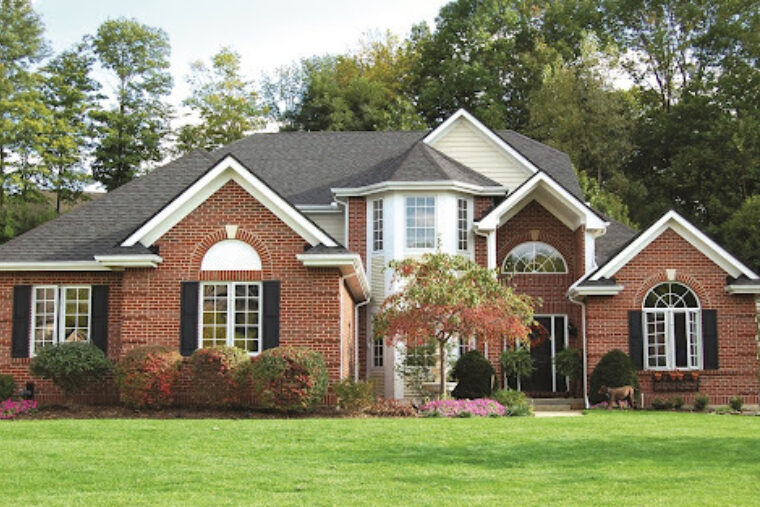 A red brick, two story home with white trim and black shutters. The front of the home is landscaped with bushes and a small tree by the front door.