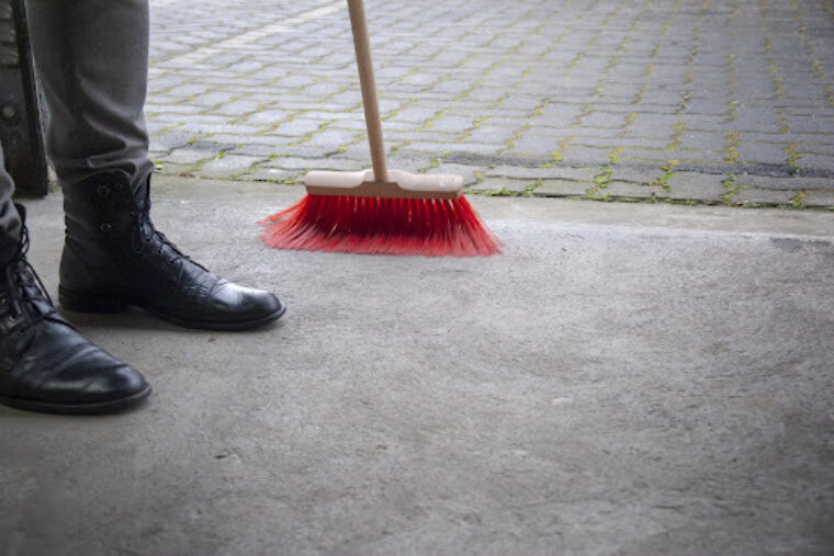 A person wearing black boots is sweeping their concrete floor with a red broom