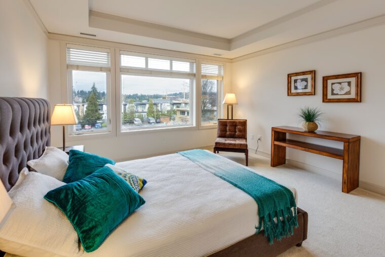 A bedroom with an empty bed decorated with green pillows and a green blanket, is shown. The room has large windows on the farthest wall, a chair and a table.